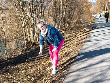 Frau in pinker Hose und Jeansjacke dehnt sich auf einem Gehweg nahe eines Flusses, umgeben von Bäumen und einem klaren Himmel an einem sonnigen Tag.