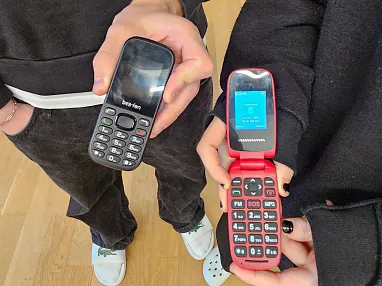 Zwei Personen halten alte Handys in der Hand: ein schwarzes Tastenhandy und ein rotes Klapphandy mit offenem Bildschirm. Sie tragen dunkle Kleidung.