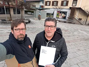 Zwei Männer mit Brillen stehen auf einem Platz in einer Stadt. Einer hält ein Blatt Papier mit Text. Im Hintergrund sind Gebäude und Schaufenster sichtbar.