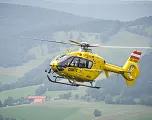 Gelber Rettungshubschrauber des ÖAMTC fliegt über eine bergige, grüne Landschaft mit Wäldern und Wiesen im Hintergrund.