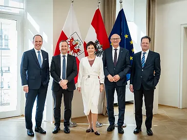 Fünf Personen in formeller Kleidung stehen lächelnd vor drei Flaggen in einem hellen Raum mit Parkettboden und Fenstern.
