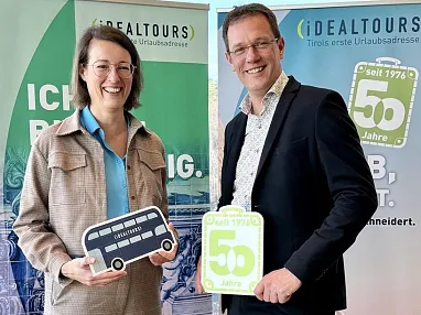 Zwei Personen halten Schilder mit der Aufschrift "50 Jahre" und "Geführte Touren" vor einem Hintergrund mit Fliesenmuster und Strandmotiv.