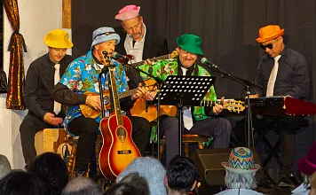 Band mit bunten Hüten und Hawaiihemden spielt live auf einer Bühne vor Publikum, mit Gitarren und Keyboard, fröhliche und entspannte Stimmung.