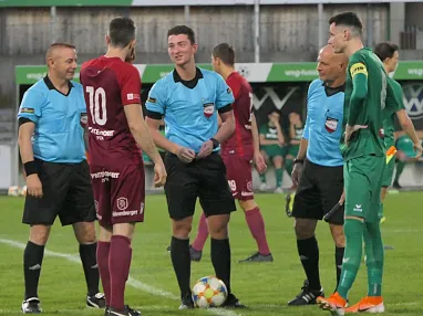 Fußballspieler und Schiedsrichter diskutieren auf einem Spielfeld. Im Hintergrund sind Werbebanner und Mitspieler zu sehen.