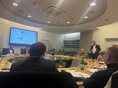 Beratungssitzung in einem modernen Konferenzraum mit ovalem Tisch, an dem mehrere Personen teilnehmen. Präsentation auf dem Bildschirm im Hintergrund.