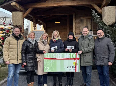 Eine Gruppe von sieben Personen steht lächelnd vor einer Holzhütte und hält ein großes, grünes Gutscheinplakat mit einer roten Schleife.