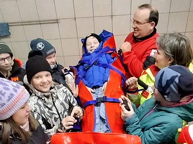 Mehrere Personen, warm gekleidet, umringen eine rote Trage in einer Rettungsübung. Einige tragen bunte Mützen, eine Person lacht. Es herrscht eine fröhliche Stimmung.