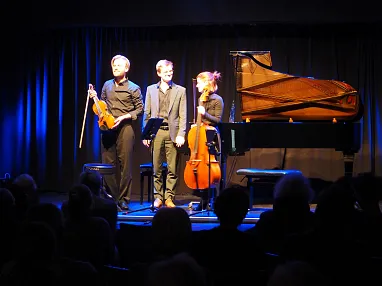Drei Musiker stehen auf einer Bühne: einer mit einer Violine, einer am Klavier und einer mit einem Cello. Der Hintergrund ist dunkel mit blauem Licht beleuchtet.