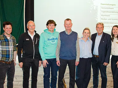 Sieben Personen stehen zusammen lächelnd vor einer Leinwand, die Teamleitungen auflistet. Eine Frau und sechs Männer, alle in Freizeitkleidung, posieren gemeinsam.