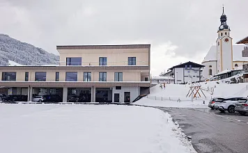 Modernes Gebäude vor schneebedeckter Landschaft und bergigem Hintergrund, neben einer traditionellen Kirche und geparkten Autos auf einem asphaltierten Weg.