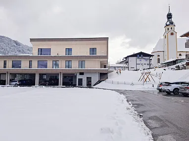 Modernes Gebäude vor schneebedeckter Landschaft und bergigem Hintergrund, neben einer traditionellen Kirche und geparkten Autos auf einem asphaltierten Weg.