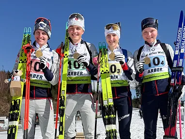 Vier Skilangläufer in sportlicher Kleidung halten Skier und Medaillen und lächeln auf einer verschneiten Piste in die Kamera.
