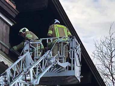 Zwei Feuerwehrleute in Schutzkleidung steigen auf einer Drehleiter zu einem Balkon eines Hauses auf, um eine Notfallmaßnahme durchzuführen.