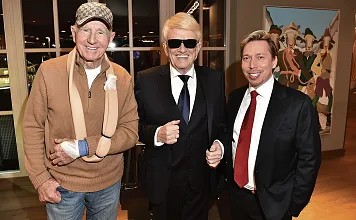 Drei Männer stehen lächelnd beisammen. Einer trägt eine Schirmmütze und Schlinge, der zweite einen Anzug mit Sonnenbrille, und der dritte ebenfalls einen Anzug.