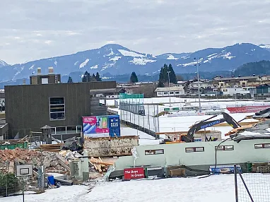 Baustelle vor schneebedeckten Bergen, mit schweren Maschinen und Gebäuden im Hintergrund. Der Himmel ist bewölkt und einige Häuser sind sichtbar.