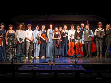 Eine Gruppe von Musikerinnen und Musikern steht auf einer Bühne, alle in eleganter Abendkleidung mit verschiedenen Musikinstrumenten wie Geigen und Cello.