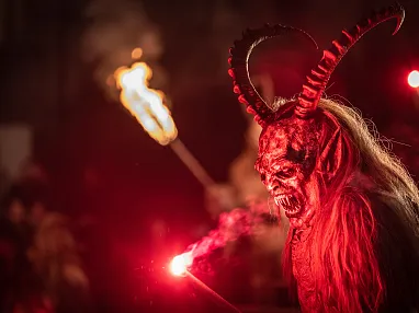 Person in gruseligem Krampus-Kostüm mit Hörnern und rotem Licht, umgeben von dunkler, festlicher Atmosphäre bei Nacht.