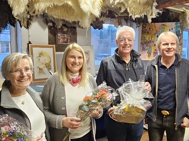 Vier Erwachsene in einem Raum mit Kunstwerken und Tierfellen, halten Geschenke in den Händen und lächeln in die Kamera. Die Atmosphäre ist freundlich und warm.