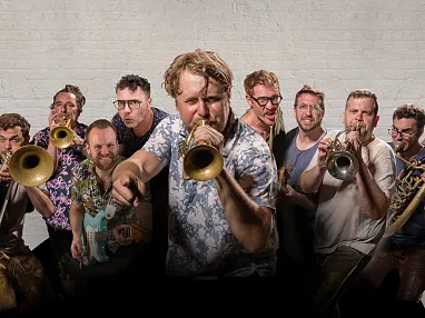 Eine fröhliche Blaskapelle mit neun Mitgliedern, die Trompeten und Posaunen spielen. Sie tragen bunte Hemden vor einem neutralen Hintergrund.