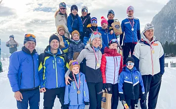 Gruppe von Menschen in Winterkleidung posiert lächelnd im Schnee. Einige tragen Medaillen. Im Hintergrund sind Berge und ein bewölkter Himmel zu sehen.