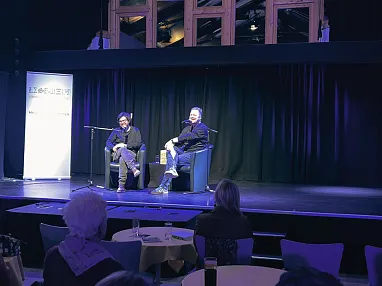 Zwei Personen sitzen auf einer Bühne in einem beleuchteten Raum, während das Publikum an Tischen sitzt und zuschaut. Die Atmosphäre ist ruhig und konzentriert.