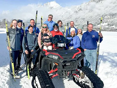 Gruppe von Menschen in Winterkleidung mit Skiern, steht zusammen im Schnee vor verschneiter Berglandschaft. Einige sitzen auf einem Fahrzeug mit Raupenantrieb.