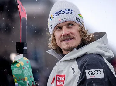Ein Mann mit blonden Locken trägt eine weiße Mütze und hält einen grünen Karton. Er hat Skier und trägt eine weiße Jacke mit mehreren Sponsorenlogos.