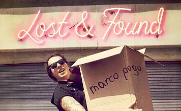 Eine Person mit Sonnenbrille und Tätowierungen hält einen großen Karton mit der Aufschrift "marco pogo" vor einem Gebäude mit neonpinkem Schriftzug.