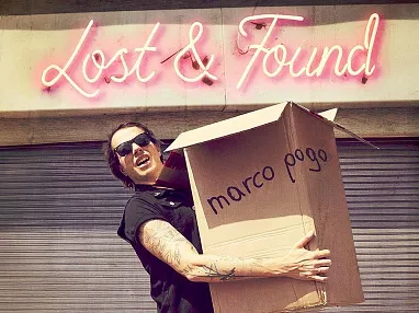 Eine Person mit Sonnenbrille und Tätowierungen hält einen großen Karton mit der Aufschrift "marco pogo" vor einem Gebäude mit neonpinkem Schriftzug.