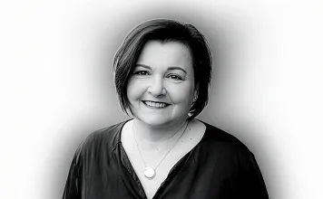 Porträt einer lächelnden Frau mit schulterlangem, dunklem Haar und einer Halskette, in einem dunklen Oberteil vor einem hellen Hintergrund.