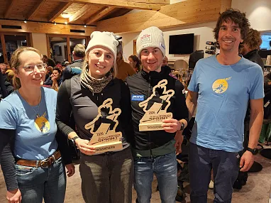 Vier Personen in Wintersportkleidung stehen zusammen, zwei halten Trophäen und lächeln in die Kamera. Im Hintergrund sind weitere Menschen zu sehen.