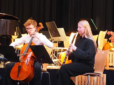 Zwei Musiker in schwarzer Kleidung spielen auf einer Bühne; eine Frau mit einer Klarinette und ein Mann am Cello, umgeben von Notenständern.