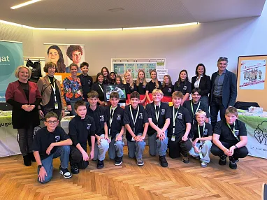 Eine Gruppe von Jugendlichen und Erwachsenen posiert in einem Raum mit Holzfußboden vor Ausstellungstischen. Einige tragen Medaillen um den Hals.