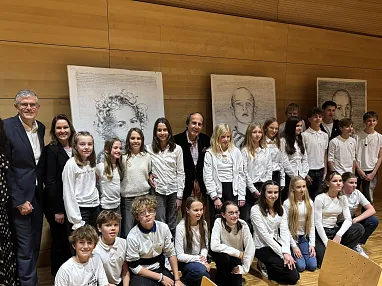 Eine Gruppe von Menschen posiert lächelnd vor Porträts an einer Holzwand. Die meisten tragen weiße Hemden. Es herrscht eine freundliche Atmosphäre.