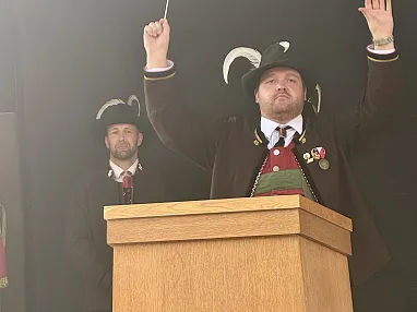 Zwei Männer in traditioneller Kleidung, einer am Rednerpult mit erhobenen Armen, der andere im Hintergrund. Beide tragen Hüte mit Federn.