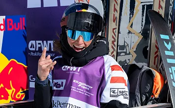 Ein Skifahrer in Winterausrüstung mit Helm und Brille zeigt lächelnd das "Rock on"-Zeichen. Im Hintergrund sind ein Red Bull-Logo und Sponsorenbanner sichtbar.