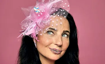 Frau mit dunklen Haaren trägt einen pinken Fascinator und ist mit Konfetti geschminkt. Sie blickt verträumt zur Seite vor rosa Hintergrund.