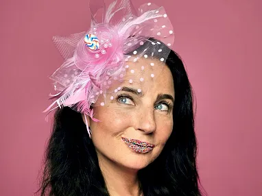 Frau mit dunklen Haaren trägt einen pinken Fascinator und ist mit Konfetti geschminkt. Sie blickt verträumt zur Seite vor rosa Hintergrund.