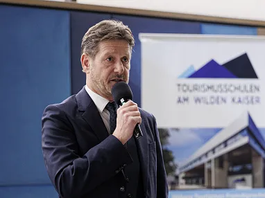 Ein Mann im Anzug hält ein Mikrofon und spricht. Im Hintergrund ist ein Banner mit der Aufschrift "Tourismusschulen am Wilden Kaiser" sichtbar.