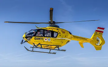 Gelber Rettungshubschrauber des ÖAMTC fliegt am klaren Himmel. Er trägt medizinische Symbole und ist in Österreich registriert.