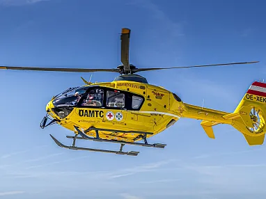 Gelber Rettungshubschrauber des ÖAMTC fliegt am klaren Himmel. Er trägt medizinische Symbole und ist in Österreich registriert.