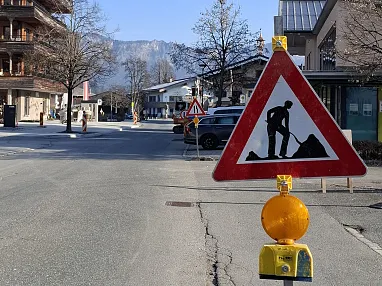 Baustellenschild auf einer leeren Straße mit Alpen im Hintergrund, sonniger Tag und einige Autos und Gebäude zu sehen.