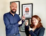 Zwei Personen lachen und zeigen auf gerahmte Auszeichnungen an einer weißen Wand. Der Mann hat einen Bart, die Frau langes Haar. Sie sind gut gekleidet.