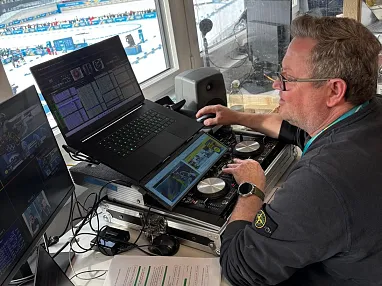 Ein Mann bedient eine DJ-Konsole mit einem Laptop und einem Tablet in einem technischen Studio. Er trägt eine Brille und ein schwarzes Oberteil.