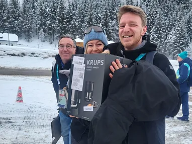 Drei Personen stehen lächelnd im Schnee, eine Person hält eine Kaffeemaschine-Box. Im Hintergrund sind verschneite Bäume und ein Verkehrskegel sichtbar.
