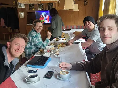 Vier Personen sitzen gemütlich an einem Tisch mit Kaffee und Laptop. Im Hintergrund läuft ein TV mit einer bunten Zeichentrickfigur.