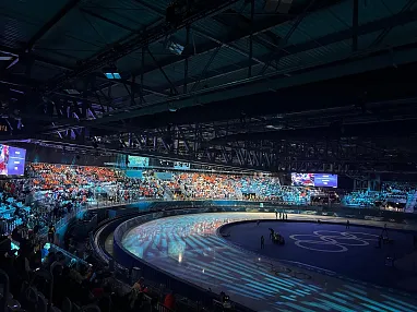 Ein Eislaufstadion mit gefüllten Tribünen. Die Eisbahn ist blau beleuchtet, das Dach hat eine industrielle Struktur. Zuschauer sind in buntem Licht getaucht.
