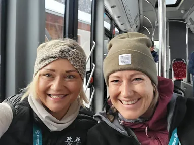 Zwei lachende Frauen in Winterkleidung sitzen in einem Bus vor einem schneebedeckten Fenster. Im Hintergrund sind leere Sitze und ein Passagier zu sehen.
