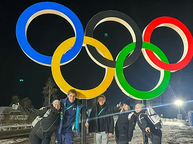 Fünf Personen posieren fröhlich bei Nacht in einem verschneiten Gebiet vor den beleuchteten Olympischen Ringen in den Farben Blau, Schwarz, Rot, Gelb und Grün.