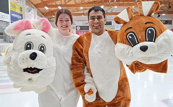 Zwei Personen in Tierkostümen stehen auf einer Eislaufbahn. Sie tragen große, auffällige Tierköpfe und lächeln in die Kamera, umgeben von Stadionbeleuchtung.
